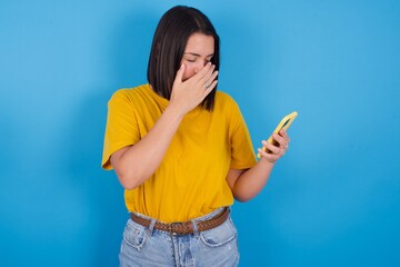 young beautiful brunette girl with short hair standing against blue background looking at smart phone feeling sad holding hand on face.