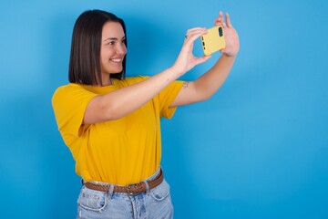 young beautiful brunette girl with short hair standing against blue background taking a selfie to post it on social media or having a video call with friends.