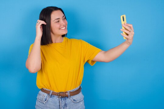 young beautiful brunette girl with short hair standing against blue background smiling and taking a selfie ready to post it on her social media.