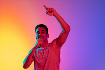 Portrait of Latina young man, singer with microphone performing isolated on gradient yellow purple background in neon light. Concept of emotions, facial expression