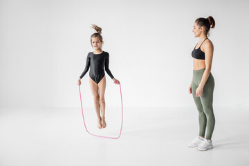 Young mom or sports coach trains little girl in rhythmic gymnastics with jumping rope at white...