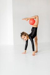 Little girl practising rhythmic gymnastic with a ball at white room. Children's gymnastics and training from an early age - obrazy, fototapety, plakaty