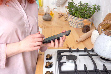 Young happy woman holding in hands a mobile phone standing by the stove on the white, light kitchen in the morning. Close up