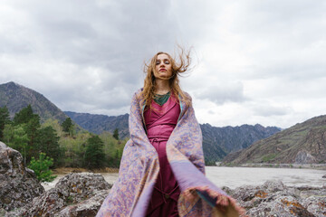 Naklejka premium Young beautiful shaman-looking woman standing in the wind in mountains wearing ethnic dress with scarf