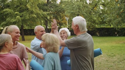 Group of cheerful senior people walking in park with exercise mats and greeting female fitness coach with hugs