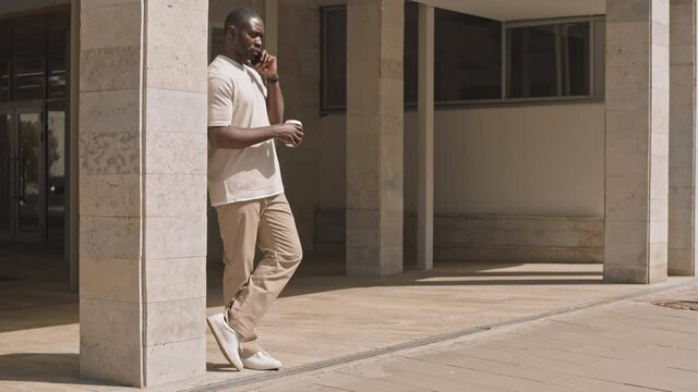 Full shot of young African-American man in casualwear standing outside modern office building with cup of coffee and making phone call arranging meeting with friend or colleague
