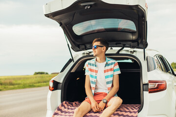 Young man in sunglasses sitting in the trunk of a car, outdoors on the road, summer vacation, travel