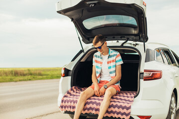 Young man in sunglasses sitting in the trunk of a car, outdoors on the road, summer vacation, travel