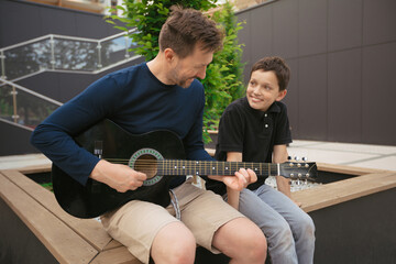Young father teaches his son to play guitar summer day off in city square .Concept of a modern...