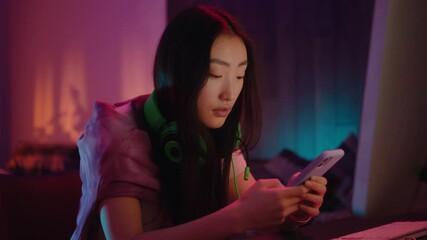 Portrait asian young woman with headphone use phone in neon light room at home. Joyful, homey, smartphone, chatting, attractive, youth, darkness, lady, alone, typing, tapping, resting. Slow motion
