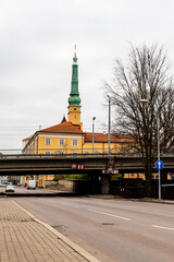 Vertical photo of street view to horizontal traffic bridge, yellow Riga’s castle and church tower. November vibe on street. Street, traffic, bridge, lamps, tree, castle, church and grey autumn sky.