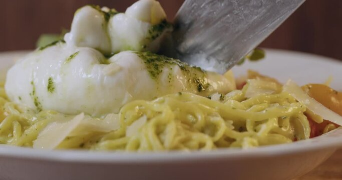 Close up of sharp knife cutting burrata cheese on freshly cooked italian pasta. Delicious main cours at modern restaurant. Enjoyment from food. 
