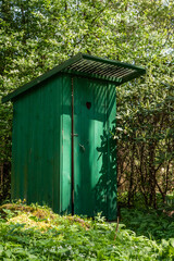 Fototapeta na wymiar Vertical green timber frame outhouse with heart shape hole in door. Rustic wooden latrine among bushes. Dry outside toilet close to green vegetation bush. Green wood jakes hided among bushes and bloom