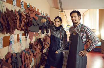 Portrait of happy staff at shoe factory. Two smiling workers and colleagues looking at camera standing near wall of peg hangers with cut out leather details. Footwear manufacturing industry concept