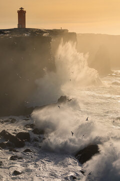 Powerful wave splashing at granite cliff with lighthouse