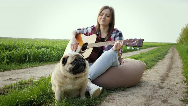 Woman Singing And Playing Guitar With Pug Dog Sitting On Bag Chair In Green Field At Sunset, Best Friends