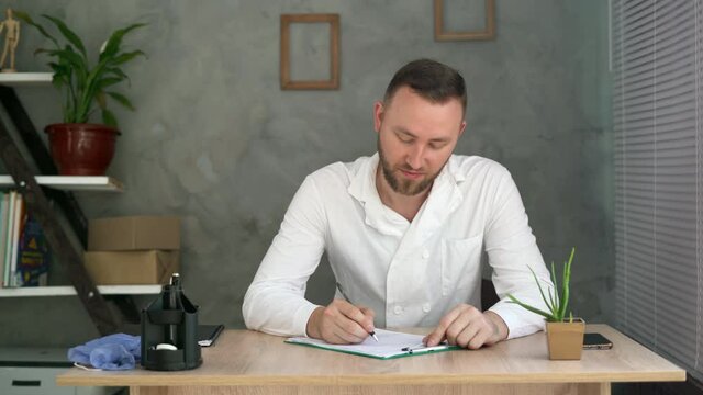 Male doctor sitting at the table and writing a report on a document in the hospital office. Medical staff and physician services.