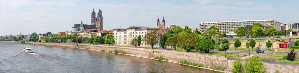 Fototapeta na wymiar Panoramic view over old cathedral at historical downtown in Magdeburg with a touristic boat coming by at Elbe River, Germany, at blue summer sky with clouds.