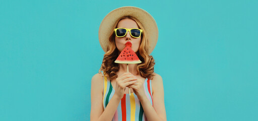 Summer portrait of stylish young woman eating a lollipop or ice cream shaped slice of watermelon wearing a straw hat on blue background