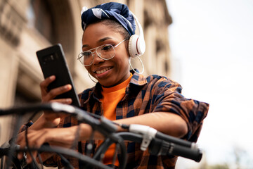 Happy young woman riding bicycle in the city. Beautiful african woman using the phone outdoors