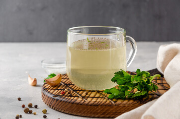 Chicken broth in a glass cup with parsley, garlic and other spices on a wooden board on a gray concrete background. Copy space.