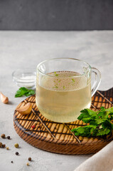 Chicken broth in a glass cup with parsley, garlic and other spices on a wooden board on a gray concrete background. Copy space.