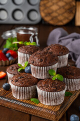 Chocolate muffins or cupcakes with chocolate drops on a wooden board on a gray background with fresh berries and mint. Copy space.