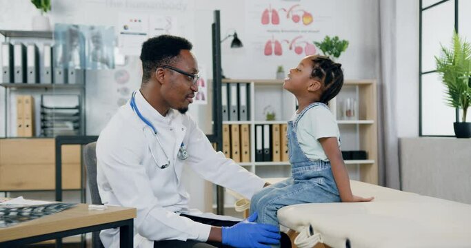 Good-looking smiling caring african american doctor talking with joyful little girl in modern medical office and giving her high five,medicine concept