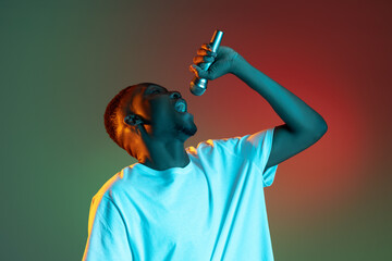 Young african man singing with microphone on neon gradient background. Concept of emotion, action, facial expression.