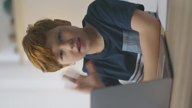 Generation Z. Little redhead boy greeting, waving hand to camera, sitting with laptop at home, video for vertical story