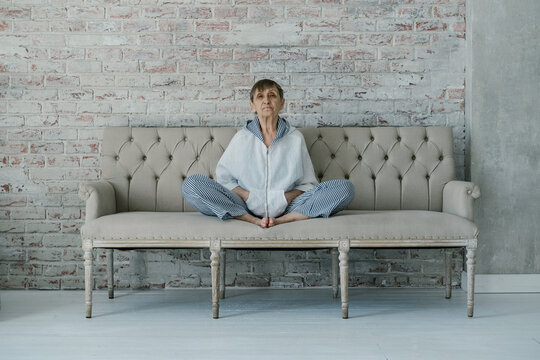 Athletic elderly woman wearing white suit sitting on sofa in lotus position