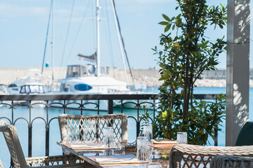 Outdoor cafe tables on the seaside embankment of mediterranean town in a summer day. Cafe offers a sea and marina view with yachts 