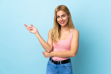 Young Uruguayan blonde woman over isolated blue background happy and pointing up