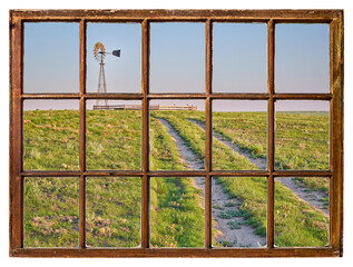 dirt road in a green prairie leading to a windmill with a water pump in shortgrass prairie as seen from a vintage sash window