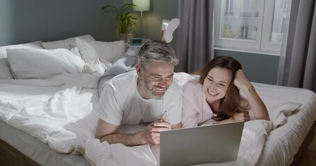 Image of romantic cheerful couple using laptop and chatting while lying on bed in evening room at home. Woman looking at her man and smiling in front of the laptop screen - Powered by Adobe