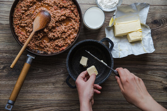 Woman cooking homemade classic lasagna bolognese, on dark table; with ingredients, top view copy space, hands in picture