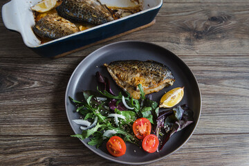 Baked butterfish or Pampanito fish with salad and lemon in a black plate with cutlery on a wooden table background. Cooking fish.