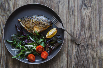 Baked butterfish or Pampanito fish with salad and lemon in a black plate with cutlery on a wooden table background. Cooking fish.