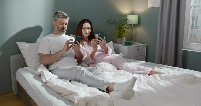 Morning with gadgets. Caucasian couple sitting at the bed at the morning and ignoring each other while using their gadgets at the free time. Internet addiction and gadgets concept