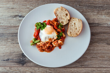 Shakshuka with a baguette on a white plate on a white wooden table background. Poached eggs in spicy tomato sauce. Summer breakfast with eggs and tomato sauce.