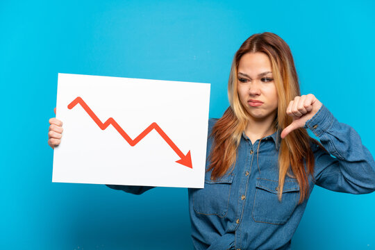 Teenager girl over isolated blue background holding a sign with a decreasing statistics arrow symbol and doing bad signal
