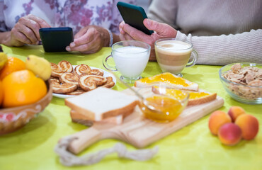Two people holding mobile phones while having breakfast at home. Close up on the table with corn flakes, cookies and marmelade