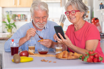 Beautiful elderly couple having breakfast at home, woman using smart phone. Senior people relaxed and happy