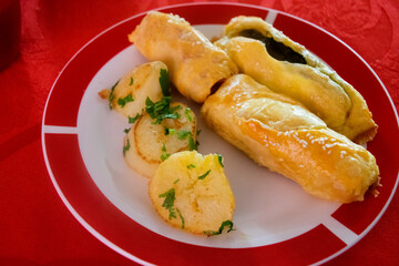 Plate served on table with red tablecloth, plate of baked rolls of spinach and ham served with potatoes seasoned with parsley.