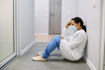 Fototapeta na wymiar Tired female doctor after a hard working day in a medical clinic sits on floor in corridor hospital with her eyes closed, wearing a medical mask. Pandemia coronavirus