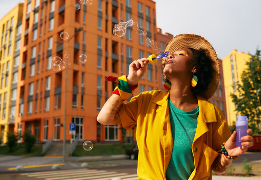 Stylish multi-ethnic woman blowing bubbles walking around modern city with colored buildings. Summer life in city