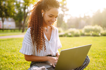 Freelancer woman using laptop computer sitting on grass at park. Young woman working online or...