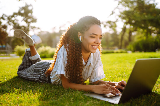 Freelancer woman using laptop computer sitting on grass at park. Young woman working online or studying and learning while using notebook. 
