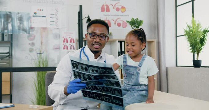 Attractive smiling caring black-skinned male doctor showing results of x-ray for little girl in modern medical center during scheduled appointment