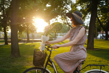 Young woman in pink dress riding a retro bicycle and holding bouquet of flowers in summer